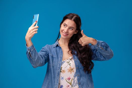 Young pretty girl in a white dress with floral print and blue denim shirt is posing with a smartphone on a blue background. She is making a selfie. People sincere emotions, lifestyle concept. Mockup copy space.