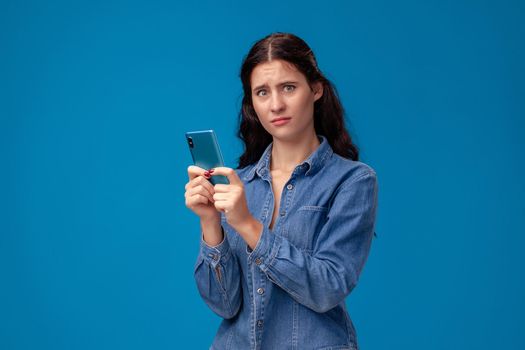 Young nice woman in a blue denim shirt is posing with a smartphone on a blue background. She is chatting with someone and looking confused. People sincere emotions, lifestyle concept. Mockup copy space.