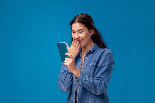 Young cheerful girl in a blue denim shirt is posing with a smartphone on a blue background. She is chatting with someone and looking wondered. People sincere emotions, lifestyle concept. Mockup copy space.