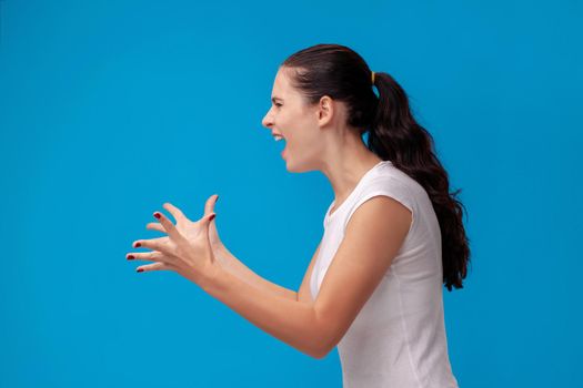 Studio portrait of a lovely girl in a white t-shirt against a blue wall background. She stands in profile, screaming at someone and gesturing. People sincere emotions, lifestyle concept. Mock up copy space.