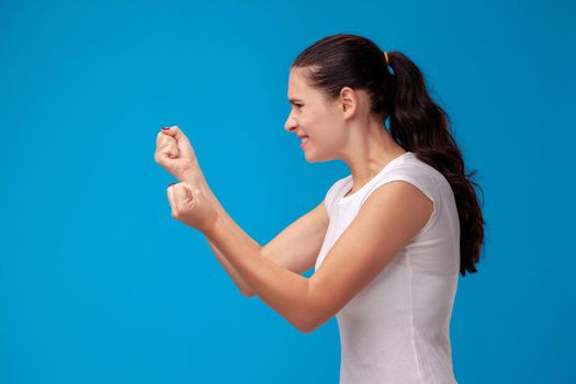 Studio portrait of a lovely female in white t-shirt against a blue wall background. Girl stands in profile and showing her fists to someone. People sincere emotions, lifestyle concept. Mock up copy space.