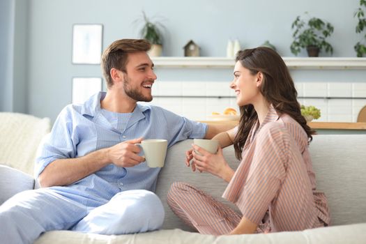 Beautiful young couple in pajamas is looking at each other and smiling on a sofa in the living room