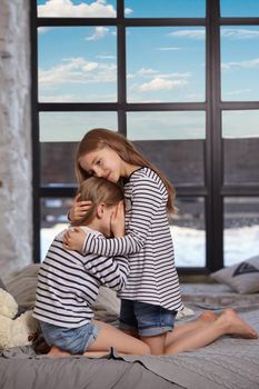 Two little sisters in striped t-shirts and denim shorts are sitting on the sofa in the bright spacious room. Little sister upset and crying. Oolder sister hugs younger and holds her head