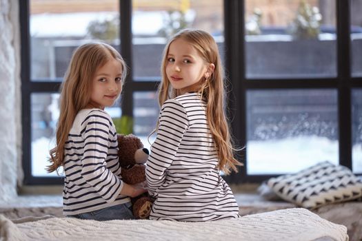 Two cute little sisters sitting on the bed in their bedroom before big window back to camera. They are looking at camera over their shoulders