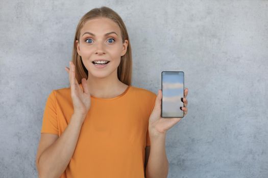 Portrait of a smiling young woman showing blank screen mobile phone isolated over grey background.