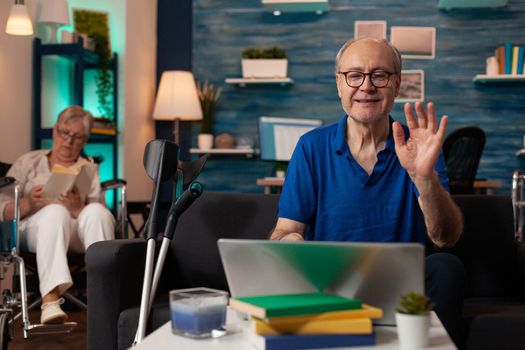 Senior man waving at video call conference on laptop at home. Older adult using online remote communication with device on couch while retired disabled woman sitting in wheelchair