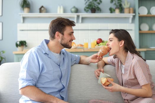 Happy young couple in pajamas in kitchen having breakfast, feeding each other a croissant