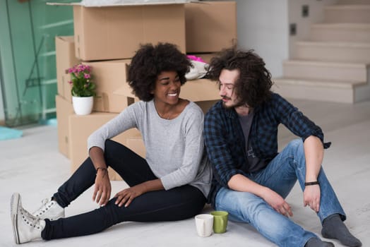 Relaxing in new house. Cheerful young multiethnic couple sitting on the floor and drinking coffee while cardboard boxes laying all around them