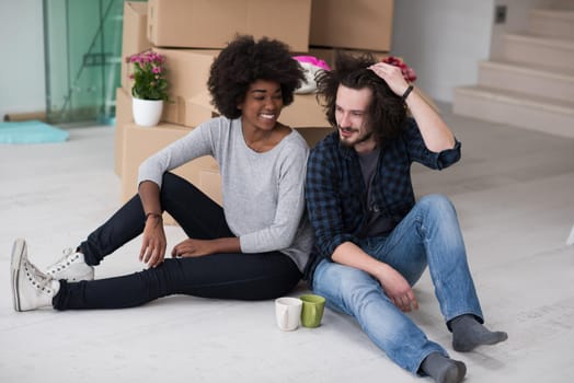 Relaxing in new house. Cheerful young multiethnic couple sitting on the floor and drinking coffee while cardboard boxes laying all around them