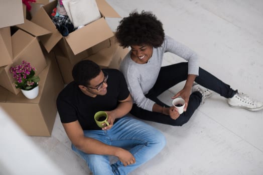 Relaxing in new house. Cheerful young African American couple sitting on the floor and drinking coffee while cardboard boxes laying all around them