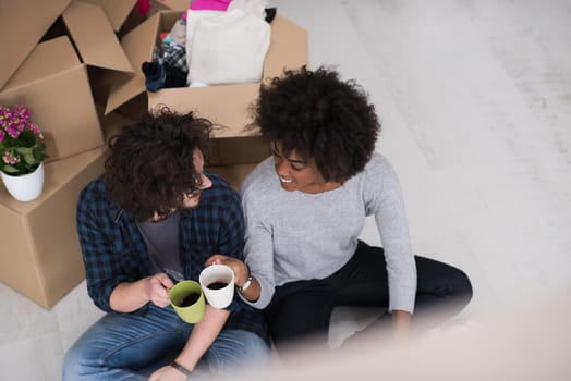 Relaxing in new house. Cheerful young multiethnic couple sitting on the floor and drinking coffee while cardboard boxes laying all around them