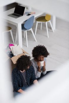 Relaxing in new house. Cheerful young multiethnic couple sitting on the floor and drinking coffee while cardboard boxes laying all around them