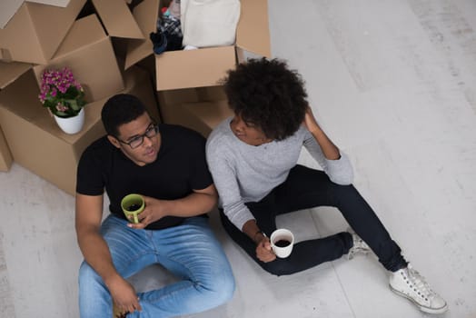 Relaxing in new house. Cheerful young African American couple sitting on the floor and drinking coffee while cardboard boxes laying all around them