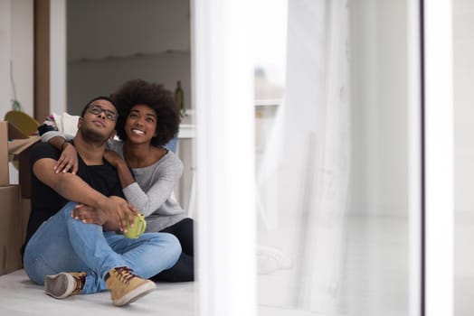 Relaxing in new house. Cheerful young African American couple sitting on the floor and drinking coffee while cardboard boxes laying all around them