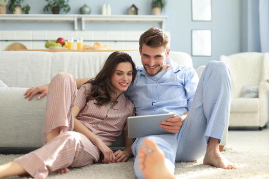 Young couple in pajamas watching media content online in a tablet sitting on the floor in the living room
