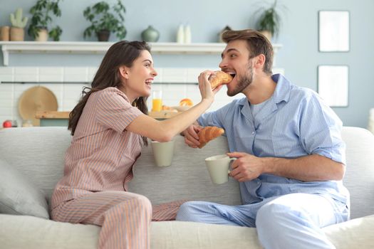 Happy young couple in pajamas in kitchen having breakfast, feeding each other a croissant