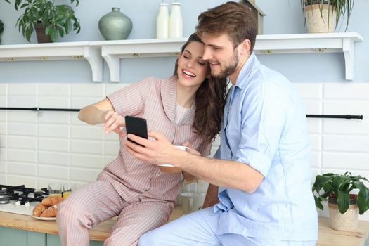 Happy young couple in pajamas watching online content in a smart phone and smiling while cooking in kitchen at home