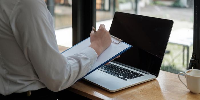 Business woman hand are taking notes on paper with a pen, and she is using a laptop computer on a wooden desk in the office.Web banner.