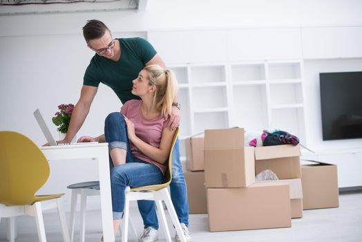 Young couple moving in a new home. Man and woman at the table using notebook laptop computer and plans with boxes around them