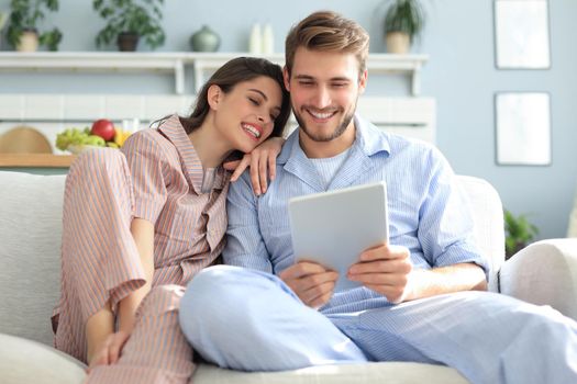 Young couple in pajamas watching media content online in a tablet sitting on a sofa in the living room