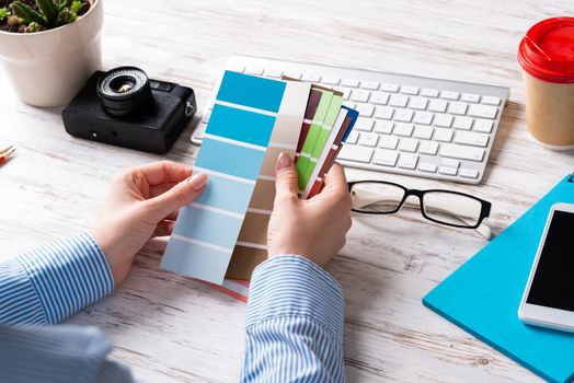 Interior designer holding color swatches at wooden desk. Office workplace with computer keyboard and camera. Brainstorming and creativity. Selection of color palette for web or product design.