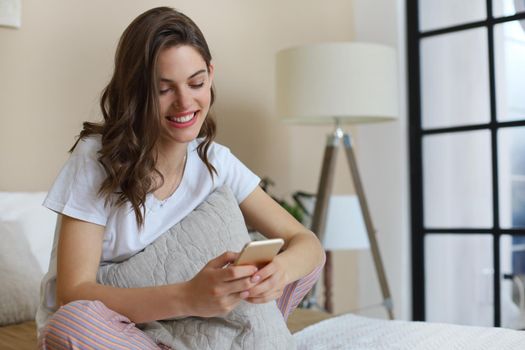 Beautiful young smiling brunette woman lying in white bed and using a phone in her bedroom.