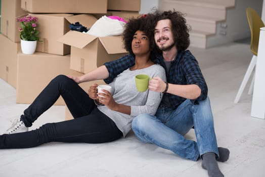 Relaxing in new house. Cheerful young multiethnic couple sitting on the floor and drinking coffee while cardboard boxes laying all around them