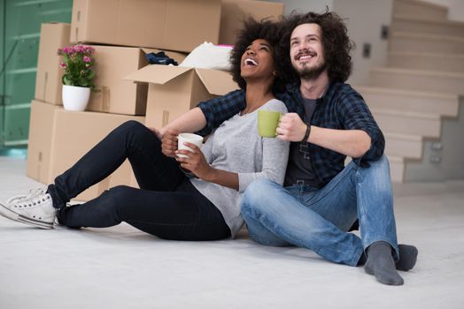 Relaxing in new house. Cheerful young multiethnic couple sitting on the floor and drinking coffee while cardboard boxes laying all around them