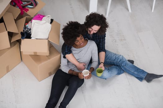 Relaxing in new house. Cheerful young multiethnic couple sitting on the floor and drinking coffee while cardboard boxes laying all around them