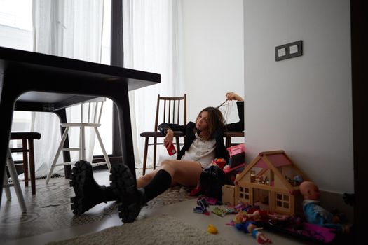 Full body childish female with smartphone touching hair and looking at camera while sitting near doll house on floor at home