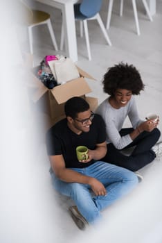 Relaxing in new house. Cheerful young African American couple sitting on the floor and drinking coffee while cardboard boxes laying all around them