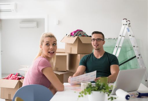 Young couple moving in a new home. Man and woman at the table using notebook laptop computer and plans with boxes around them