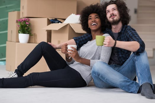 Relaxing in new house. Cheerful young multiethnic couple sitting on the floor and drinking coffee while cardboard boxes laying all around them