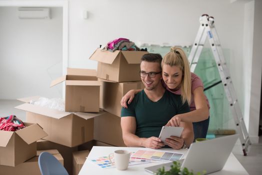 Young couple moving in a new home. Man and woman at the table using notebook laptop computer and plans with boxes around them