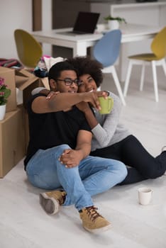 Relaxing in new house. Cheerful young African American couple sitting on the floor and drinking coffee while cardboard boxes laying all around them