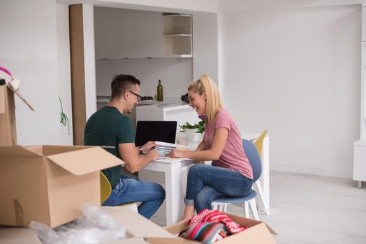 Young couple moving in a new home. Man and woman at the table using notebook laptop computer and plans with boxes around them