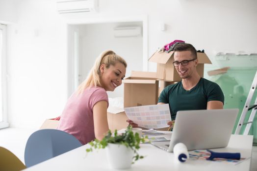 Young couple moving in a new home. Man and woman at the table using notebook laptop computer and plans with boxes around them