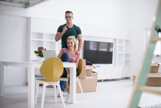 Young couple moving in a new home. Man and woman at the table using notebook laptop computer and plans with boxes around them