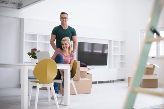 Young couple moving in a new home. Man and woman at the table using notebook laptop computer and plans with boxes around them