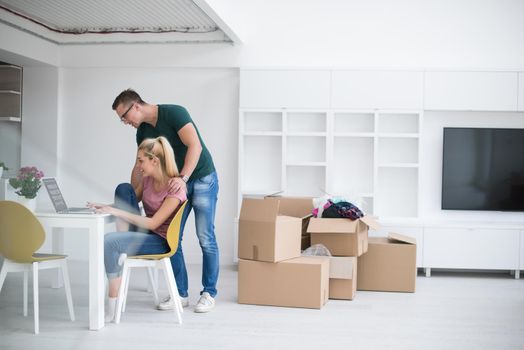 Young couple moving in a new home. Man and woman at the table using notebook laptop computer and plans with boxes around them