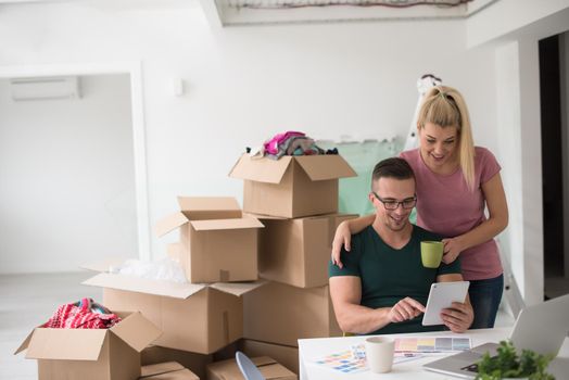 Young couple moving in a new home. Man and woman at the table using notebook laptop computer and plans with boxes around them