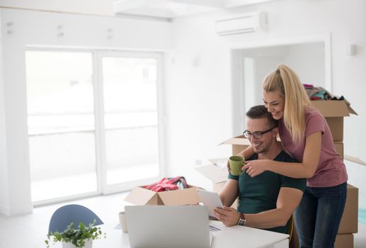 Young couple moving in a new home. Man and woman at the table using notebook laptop computer and plans with boxes around them