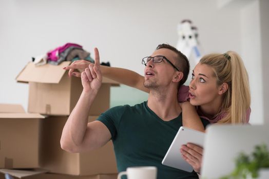 Young couple moving in a new home. Man and woman at the table using notebook laptop computer and plans with boxes around them