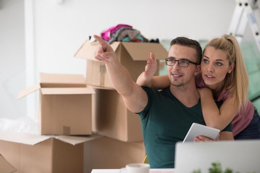 Young couple moving in a new home. Man and woman at the table using notebook laptop computer and plans with boxes around them