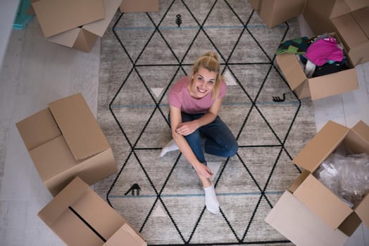 top view of young beautiful woman sitting and relaxing on the floor after moving into a new home with cardboard boxes around her