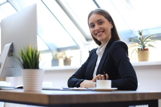 Portrait of a cheerful young businesswoman sitting at the table in office and looking at camera