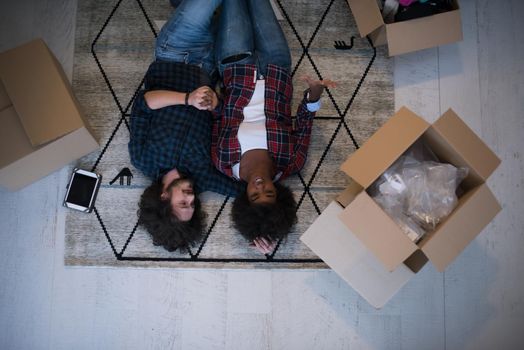 Top view of attractive young multiethnic couple moving, holding hands, looking at camera and smiling while lying among cardboard boxes