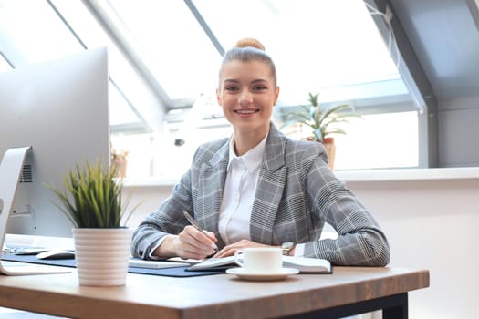 Portrait of a cheerful young businesswoman sitting at the table in office and looking at camera