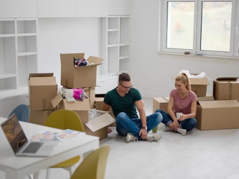 Relaxing in new house. Cheerful young couple sitting on the floor and drinking coffee while cardboard boxes laying all around them