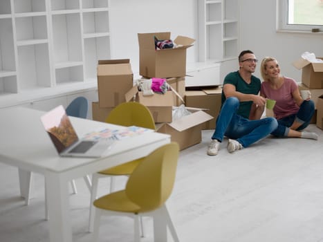 Relaxing in new house. Cheerful young couple sitting on the floor and drinking coffee while cardboard boxes laying all around them